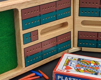 Puede incluir: Un tablero de cribbage de madera con secciones de puntuación marrones y verde azulado, un interior de fieltro verde y una baraja de cartas. Incluye clavijas naranjas y azules. La baraja de cartas está etiquetada como "PLASTIC COATED".