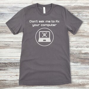 May include: A dark grey t-shirt with the text "Don't ask me to fix your computer" and a white icon of a laptop with a skull and crossbones inside.