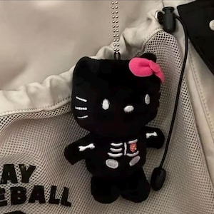 Op de afbeelding: Zwart pluche Hello Kitty speelgoed met een skeletontwerp en een roze strik. Het speelgoed hangt aan een zilveren ketting.