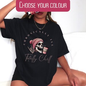 May include: Black t-shirt featuring a graphic of a pink-hatted skull holding a coffee cup. The text "Chronically Ill Totally Chill" is printed on the shirt.