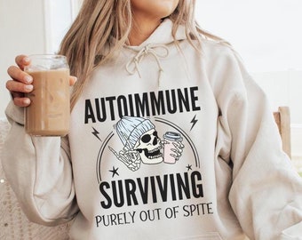 May include: A white hoodie with a black graphic of a skeleton wearing a blue beanie and holding a coffee cup. The text on the hoodie reads "Autoimmune Surviving Purely Out of Spite."