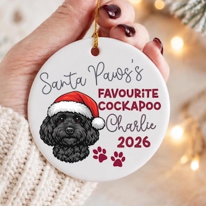 May include: A round white ceramic ornament featuring a black Cockapoo dog wearing a red and white Santa hat. The text reads Santa Paws's FAVOURITE COCKAPOO Charlie 2026, accompanied by two small red paw prints. A gold string is attached to the top.