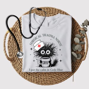 Puede incluir: Camiseta blanca con un dibujo animado en blanco y negro de una criatura peluda con un gorro de enfermera y sosteniendo una taza de café. El texto "Surgical Trauma Nurse" está por encima del dibujo animado y "I put the calm in Code Blue" está por debajo del dibujo animado.