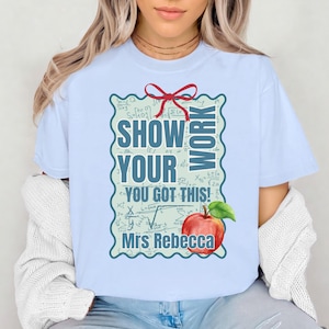 May include: Pale blue t-shirt with a graphic that reads "SHOW YOUR WORK" and "YOU GOT THIS!" within a decorative frame. An apple graphic is at the bottom, with the name "Mrs Rebecca" below it. The shirt is a light blue colour.