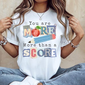 May include: White t-shirt with the text "You are MORE! More than a SCORE" in colorful block letters. The design includes an apple, a pencil, and a paper clip. The shirt is a casual style.