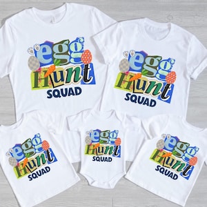 Puede incluir: Camisetas blancas y un body con un gráfico colorido de "Egg Hunt Squad". El diseño incluye las palabras "egg hunt" en azul y verde, con ilustraciones de huevos de Pascua y zanahorias. Un divertido atuendo familiar de Pascua.