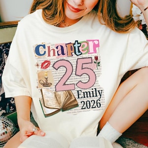 May include: A cream-colored t-shirt featuring a graphic print with the text "Chapter 25", "Emily", and "2026". The design includes a large number 25, a red lipstick kiss mark, a candle in a holder, a rose, and an open book over script text.