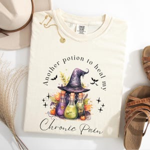Puede incluir: Una camiseta de color crema con una bruja que lleva un sombrero morado y sostiene tres frascos de poción. El texto dice "Another potion to heal my chronic pain".