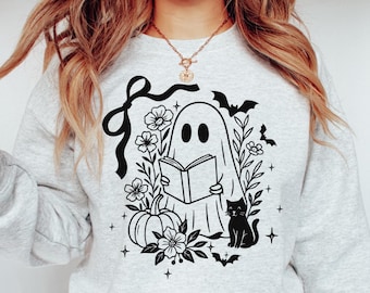 May include: Light gray sweatshirt with a black graphic of a ghost reading a book, surrounded by flowers, a pumpkin, a black cat, bats, and stars. A black bow is at the top left of the design. A Halloween-themed design.