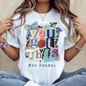 Könnte beinhalten: Weißes T-Shirt mit einem farbenfrohen Aufdruck mit der Aufschrift "You Got This". Das Design beinhaltet einen Apfel, Bleistifte und ein Papierflugzeug. Der Text "Mrs Kendal" ist darunter gedruckt.