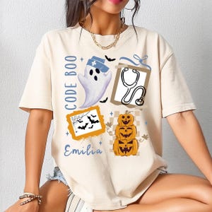 May include: A cream-coloured t-shirt featuring a Halloween design. The graphic includes a ghost in a nurse's cap, a stethoscope, stacked jack-o'-lanterns, and bats. The text "CODE BOO" and the name "Emilia" are also present.