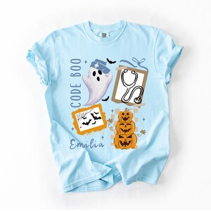 May include: Pale blue t-shirt featuring a Halloween design. The graphic includes a ghost in a nurse's cap, a stethoscope, bats, pumpkins, and the words "CODE BOO" and "Emilia".