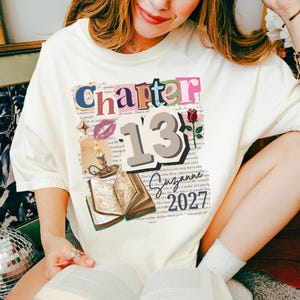 May include: A cream-colored t-shirt featuring a graphic print with the text Chapter 13 and 2027. The design includes a large number 13, a red rose, a lit candle in a holder, a purple lip print, a star, and an open book over a background of script text.