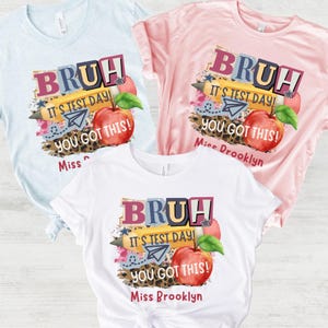 May include: Three t-shirts in light blue, pink, and white, each featuring a colorful graphic. The design includes the word "BRUH", the phrase "IT'S TEST DAY!", and an apple illustration. The text "YOU GOT THIS!" is also present.