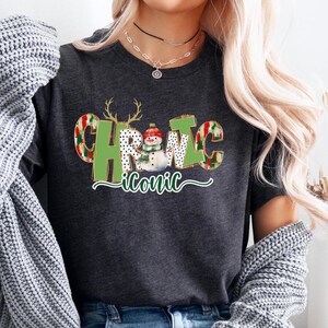 Könnte beinhalten: Ein dunkelgraues T-Shirt mit einer grünen und roten Grafik, auf der "Christmas Iconic" steht, mit einem Schneemann in der Mitte.
