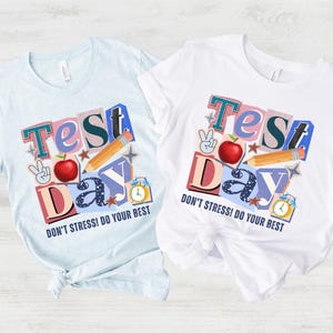 May include: Two t-shirts, one light blue and one white, each with a colorful graphic that reads "Test Day" with an apple, pencil, peace sign, and clock. The text "DON'T STRESS! DO YOUR BEST" is below the graphic.