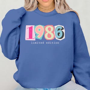 May include: A blue sweatshirt featuring the year "1986" in colourful block letters. The text "limited edition" is below the numbers. The sweatshirt is a solid blue colour.