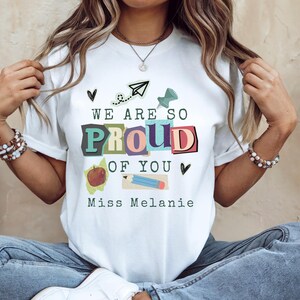 May include: White t-shirt with the text "WE ARE SO PROUD OF YOU" in colorful block letters. The shirt also features an apple, pencil, paper airplane, and push pin graphic, with the name "Miss Melanie" below.