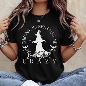 Puede incluir: Camiseta negra con un gráfico blanco de una bruja sosteniendo un arma y el texto "Chronic Illness Has Me Bat Shit Crazy".