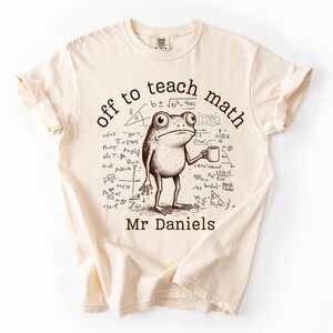 May include: A cream-coloured t-shirt with a cartoon frog holding a mug, surrounded by mathematical equations. The text reads "off to teach math" above the frog, with "Mr Daniels" below. A fun, educational design.