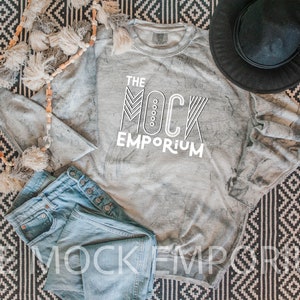 Może przedstawiać: Szara koszula z długim rękawem z białym nadrukiem "THE MOCK EMPORIUM" na czarno-białym wzorzystym dywanie, z niebieskimi dżinsami i czarnym kapeluszem.