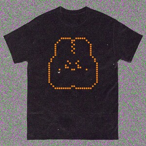 May include: Black t-shirt with an orange pixelated face design. The graphic is made up of small squares, giving a retro, digital look. The t-shirt has a classic crew neck, suitable for everyday wear.