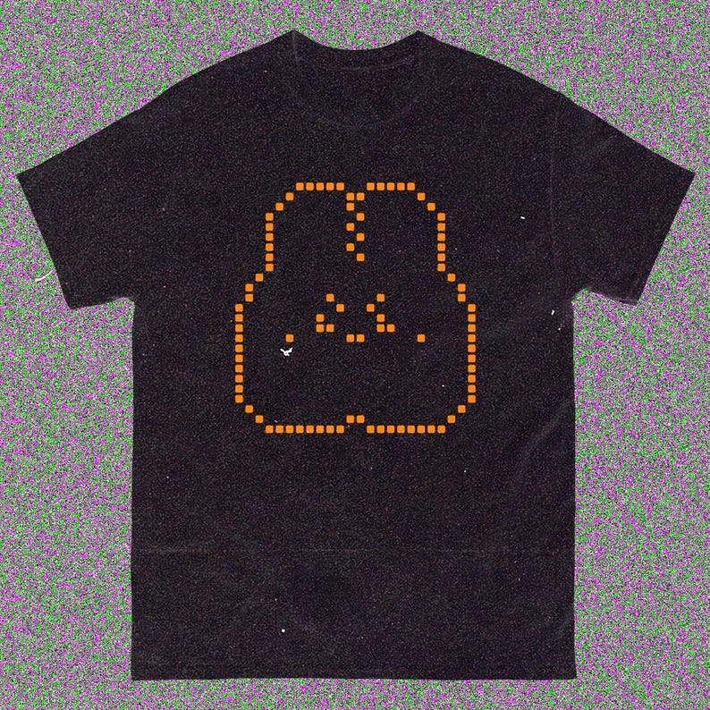 May include: Black t-shirt with an orange pixelated face design. The graphic is made up of small squares, giving a retro, digital look. The t-shirt has a classic crew neck, suitable for everyday wear.