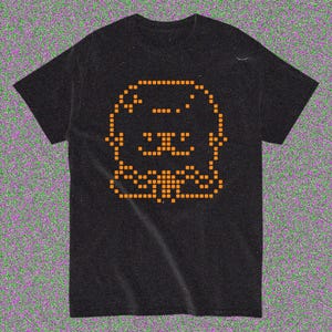 May include: Black t-shirt featuring an orange pixelated graphic of a cartoon animal face. The design is centered on the front of the shirt. The shirt has short sleeves and a crew neck.