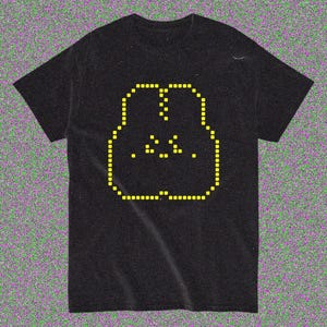 May include: Black t-shirt with a yellow pixelated face design. The face is outlined with small yellow squares, with yellow dots for the eyes and mouth. The shirt is made of a soft material.