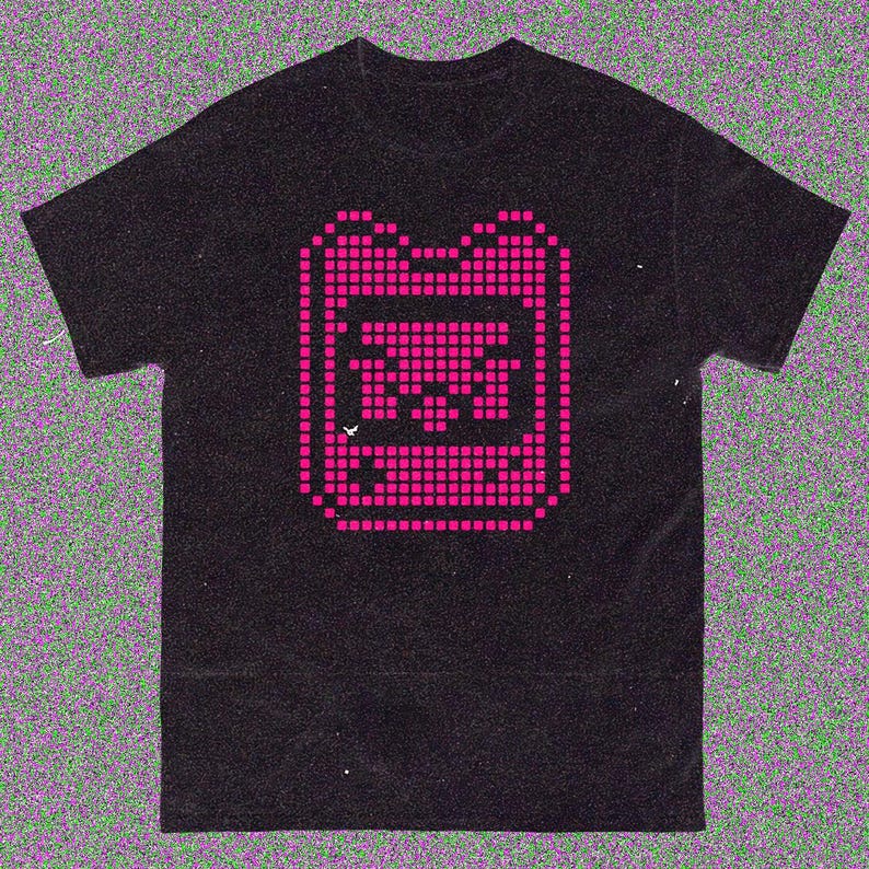 May include: Black t-shirt with a pixelated pink graphic of a retro handheld gaming device. The design is centered on the shirt and is made up of small, square pixels. The t-shirt has a classic crew neck.