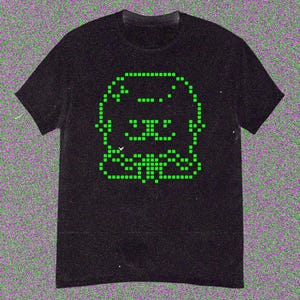 May include: Black t-shirt featuring a pixelated green graphic of a smiling creature. The design is made up of small, square pixels, giving it a retro video game aesthetic. The shirt has short sleeves and a crew neck.