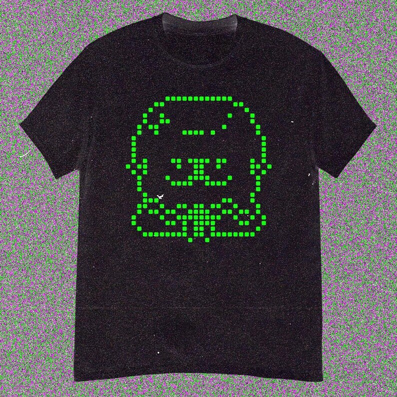May include: Black t-shirt featuring a pixelated green graphic of a smiling creature. The design is made up of small, square pixels, giving it a retro video game aesthetic. The shirt has short sleeves and a crew neck.