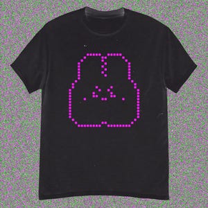 May include: Black t-shirt with a pixelated pink face design. The graphic is formed from small squares, giving a retro digital look. The tee has a crew neck and short sleeves, suitable for everyday wear. The design is centered on the chest.