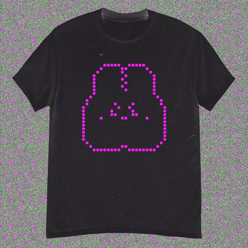 May include: Black t-shirt with a pixelated pink face design. The graphic is formed from small squares, giving a retro digital look. The tee has a crew neck and short sleeves, suitable for everyday wear. The design is centered on the chest.
