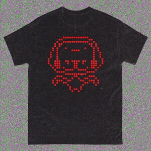 May include: Black t-shirt with a red pixelated dog graphic. The dog wears headphones and a bow tie. The design is created from small squares, giving a retro video game feel. The shirt is made of a soft material.