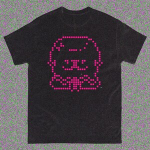 May include: Black t-shirt featuring a pixelated pink graphic of a cartoon animal. The design is centered on the front of the shirt. The t-shirt has a crew neck and short sleeves.