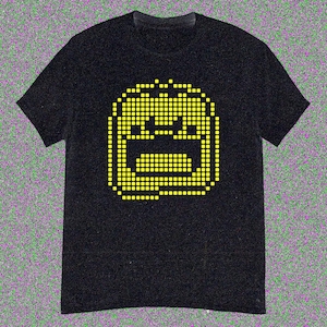 Puede incluir: Camiseta negra con un diseño de cara amarilla pixelada. El gráfico muestra una cara cuadrada con ojos, nariz y boca. La camiseta tiene mangas cortas y cuello redondo.