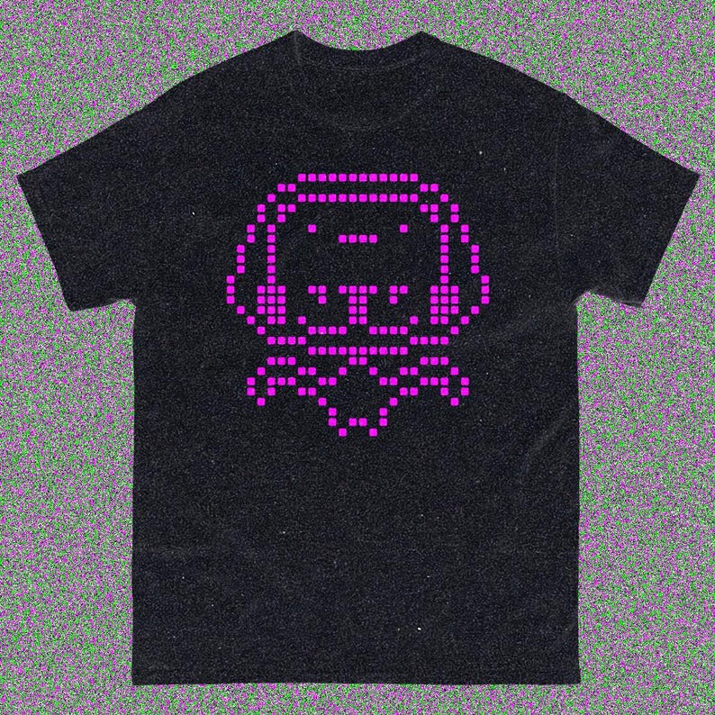 May include: Black t-shirt with a pixelated pink dog design. The dog has a square head, ears, and a bow tie. The design is made up of small squares, giving it a retro, digital look. The shirt is made of a soft material.