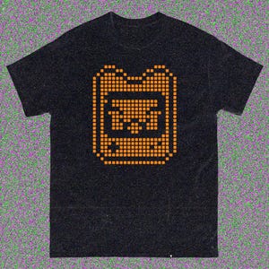 May include: Black t-shirt with an orange pixelated graphic. The graphic is a stylized design of a rectangular shape with a central image. The shirt has short sleeves and a crew neck.