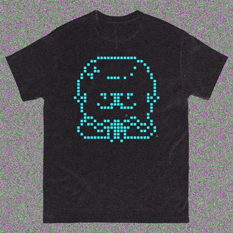 May include: Black t-shirt featuring a pixelated teal graphic of a smiling creature. The design is centered on the shirt and is made up of small squares. The shirt has short sleeves and a crew neck.