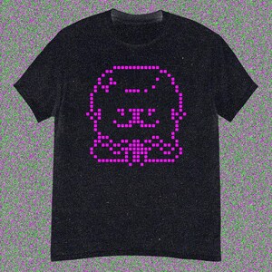 May include: Black t-shirt featuring a pixelated pink cat design. The cat's face and body are formed by small, square pixels, creating a retro aesthetic. The shirt has short sleeves and a crew neck.