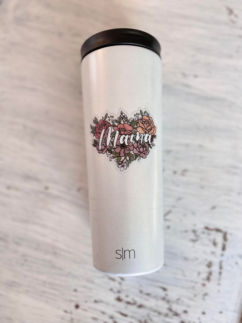 May include: White stainless steel tumbler with a floral design and the word "Mama" in pink lettering. The tumbler has a black lid and the word "slim" printed on the bottom.