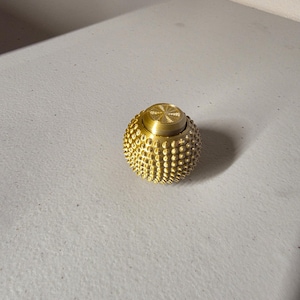 Puede incluir: Un objeto esférico dorado con una superficie texturizada de pequeñas protuberancias redondeadas. La parte superior de la esfera tiene un diseño circular. El objeto está sobre una superficie de color claro.