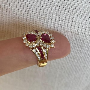 Op de afbeelding: Een gouden ring met twee opvallende peervormige rode robijnen, omringd door een cluster van kleine, heldere, rond geslepen witte diamanten. De ringband is gemaakt van gepolijst geelgoud en bevat extra kleine rode robijnen en diamanten.
