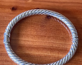 May include: A silver-colored bracelet with a twisted rope design. The bracelet is circular and has a clasp closure. The jewelry is displayed on a wooden surface.