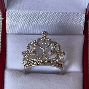May include: A silver crown ring with clear cubic zirconia stones. The ring is in a red velvet ring box.