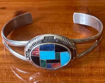 May include: A silver cuff bracelet featuring a central oval inlay of geometric stone segments in shades of turquoise, black, blue, red, and green. The metal frame has etched line details on the sides and a split band design for the wrist.