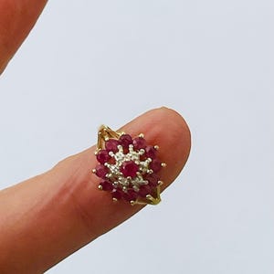Puede incluir: Un anillo de oro con un grupo de pequeñas piedras preciosas rojas ovaladas y un grupo central de piedras preciosas claras. El anillo tiene un diseño decorativo en forma de hoja en los laterales.