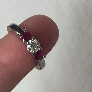 May include: A silver-toned metal ring featuring a three-stone setting. The center stone is a clear, round-cut gemstone flanked by two smaller, round-cut red gemstones. The stones are held in place by metal prongs on a smooth, polished band.