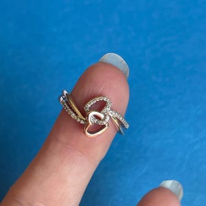 Può includere: Un anello d'argento con un accento a forma di cuore dorato e piccole gemme trasparenti. L'anello ha un design delicato con due fasce intrecciate e un singolo cuore. L'anello è mostrato su uno sfondo blu.